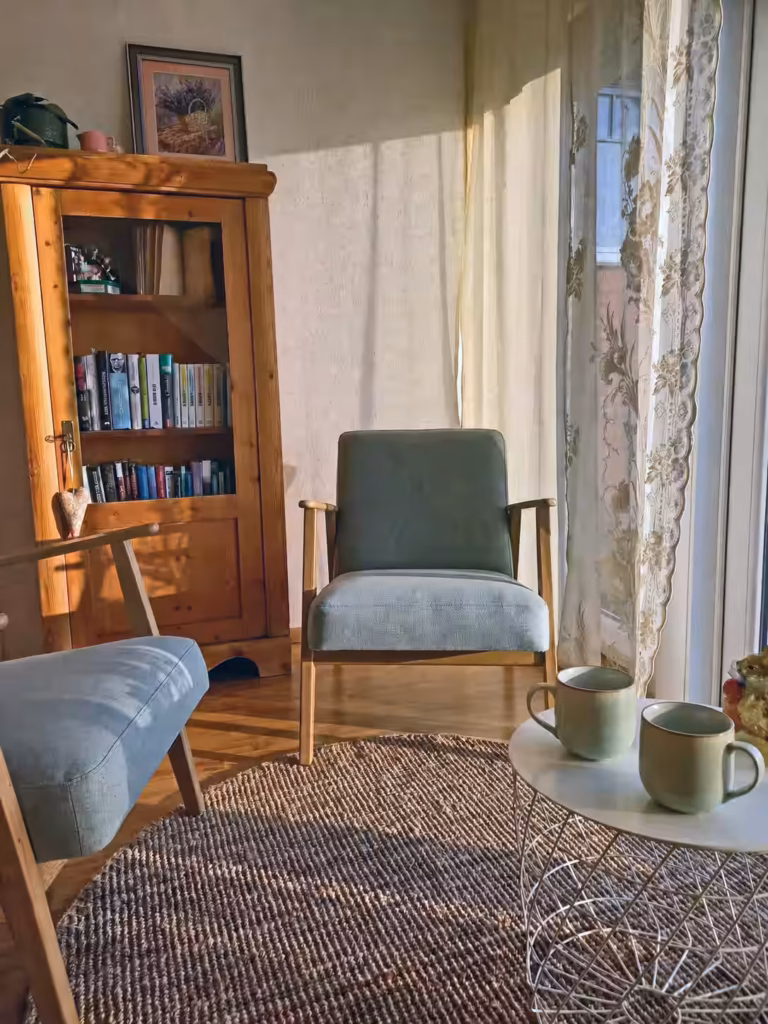 Sunlit therapy room with 2 chairs and table with 2 sage green mugs. Bookshelf in the background.